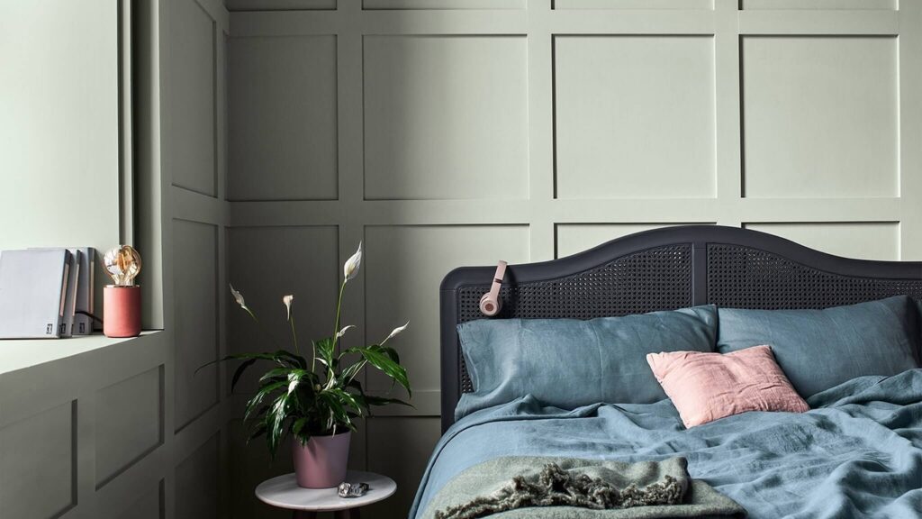 Sovrum med ljusgrå väggar i sektionerad paneldesign, mörkgrå sänggavel i rotting, blå sängkläder och en rosa kudde. På ett litet runt bord står en grön växt i rosa kruka.